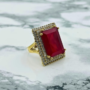 May include: A gold ring featuring a large, red, rectangular gemstone set within a border of smaller, clear gemstones.