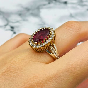 Peut inclure: Une bague en argent avec une grosse pierre rouge entourée de petites pierres claires. La bague a une bande dorée.
