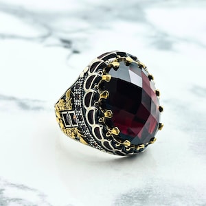 May include: A silver ring with a large, faceted, dark red gemstone set in a gold-toned crown-like setting. The ring has intricate details and a black enamel finish.