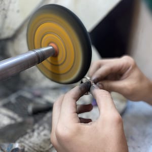 May include: A person's hand holding a silver ring against a spinning buffing wheel with a yellow and black surface.