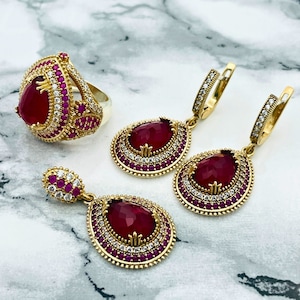 May include: A set of gold-tone jewellery with red gemstone accents. The set includes a ring, a pair of earrings, and a pendant. The jewellery is decorated with small white stones.