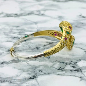 May include: A gold and silver bangle bracelet with a snake design. The snake is adorned with green and red gemstones and small diamonds.