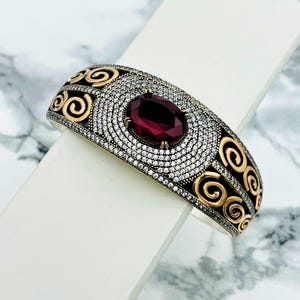 Peut inclure: Un bracelet jonc en argent et or avec une pierre précieuse rouge et un motif en spirale. Le bracelet est serti de pierres de zircone cubique transparentes.
