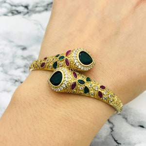 May include: A gold-toned snake-shaped bangle bracelet with green and pink gemstones. The bracelet is adorned with intricate details and features two emerald-green stones.