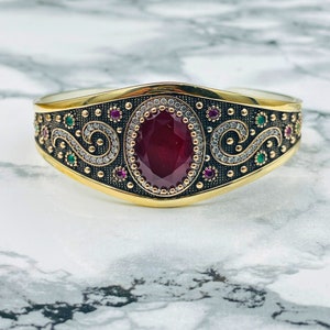 May include: A gold and silver bangle bracelet featuring a large red gemstone surrounded by smaller white gemstones. The bracelet is adorned with intricate swirls and green and pink gemstones.