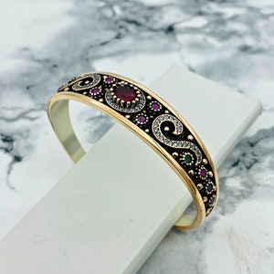 Peut inclure: Un bracelet jonc en or avec un motif en émail noir en forme de volutes et de petites pierres précieuses rouges et vertes. Le bracelet a une seule pierre rouge plus grande au centre.