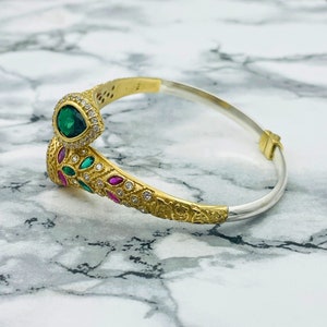 May include: A gold and silver bangle bracelet with a green and pink gemstone design. The bracelet is decorated with intricate details and features a pear-shaped green gemstone.