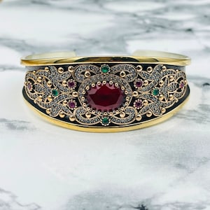 May include: A silver and gold bracelet with a large red gemstone surrounded by smaller green and red gemstones. The bracelet is decorated with intricate filigree designs.