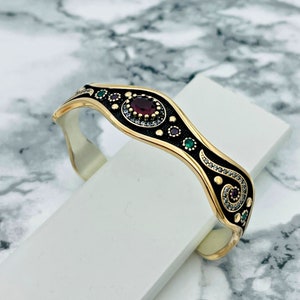 May include: A gold-coloured cuff bracelet with a black enamel inlay. The bracelet features a central oval-shaped red gemstone, surrounded by smaller gemstones in various colours. The design includes decorative swirls and dots.