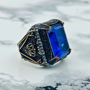 May include: A silver ring with a large, dark blue gemstone set in a detailed, ornate design. The ring has a black and gold finish with intricate details.
