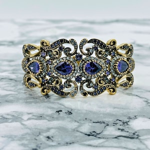May include: A gold-toned bracelet with an intricate design featuring purple and clear stones. The bracelet has a filigree pattern and is adorned with multiple marquise-shaped stones.