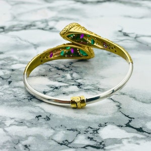 May include: A gold and silver snake bangle bracelet with green and pink gemstones. The bracelet is open and has a hinged clasp.