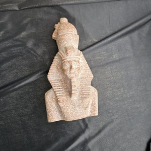 May include: A small, tan-colored bust of an Egyptian pharaoh, featuring a detailed headdress and beard. The sculpture is made of a stone-like material and is set against a dark background. The bust is a representation of ancient Egyptian art.