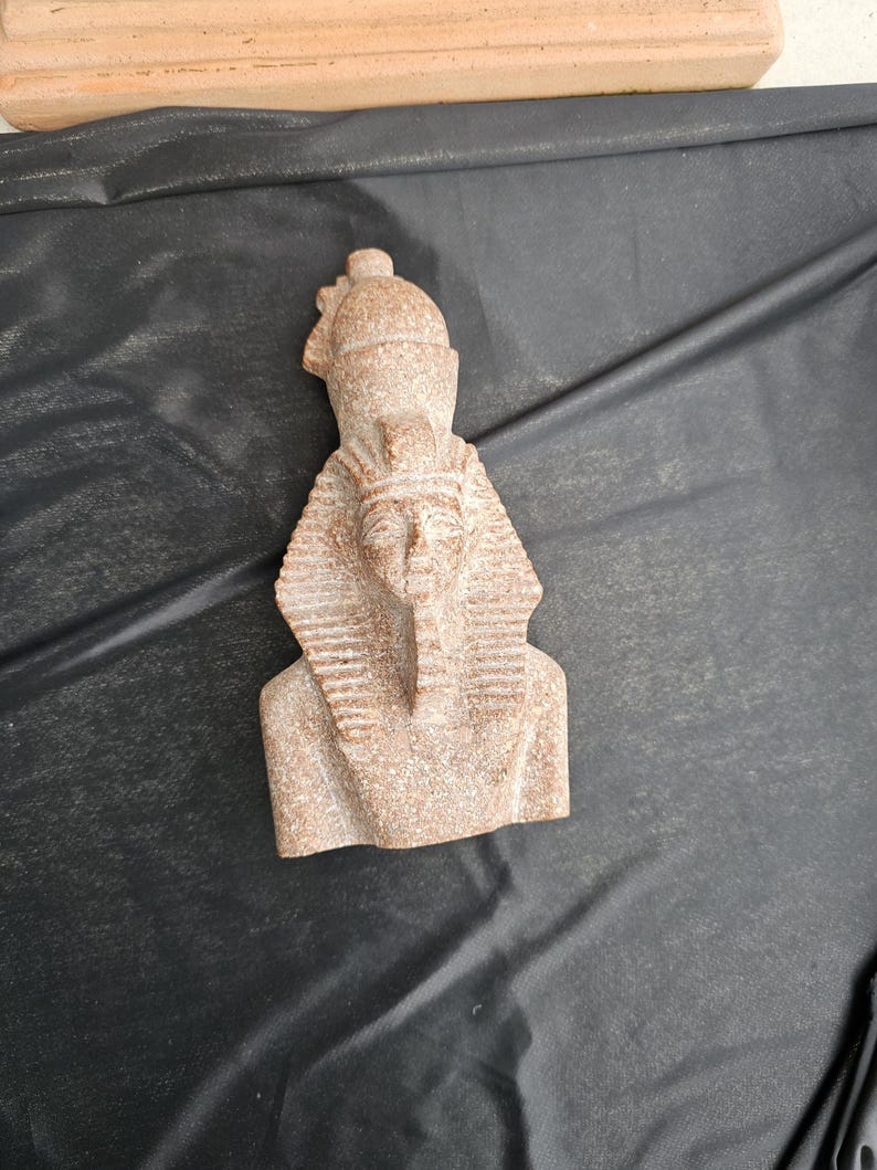 May include: A small, tan-colored bust of an Egyptian pharaoh, featuring a detailed headdress and beard. The sculpture is made of a stone-like material and is set against a dark background. The bust is a representation of ancient Egyptian art.