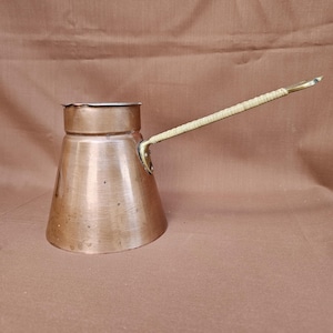 Puede incluir: Una cafetera de cobre con un mango largo y tejido. La cafetera tiene forma cónica con una base ancha y una parte superior estrecha. El mango está unido al lateral de la cafetera y se curva hacia arriba. La cafetera es de cobre y tiene un acabado brillante.