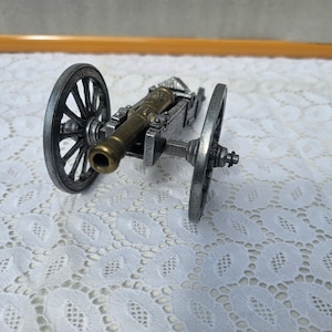 Könnte beinhalten: Ein Miniaturkanonenmodell mit einem Bronzelauf und silberfarbenem Wagen und Rädern. Die Räder haben ein Speichendesign. Die Kanone steht auf einer weißen Spitzentischdecke.