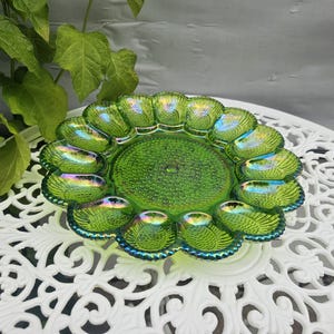May include: A round, vibrant green glass egg plate featuring an iridescent finish and a textured, stippled center. The plate has twelve individual, shell-shaped indentations arranged in a circular pattern around the perimeter with a scalloped edge.