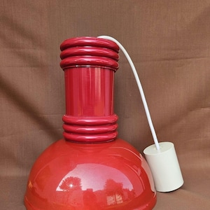 Può includere: Lampada a sospensione rossa con finitura lucida. La lampada ha una parte superiore cilindrica con dettagli a coste, una base arrotondata e un cavo bianco con un tirante cilindrico. Progettata per l'installazione a soffitto.