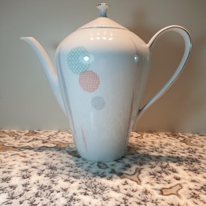 May include: White ceramic coffee pot with a lid and a handle. The pot has a geometric pattern of circles in shades of green, pink, and gray.