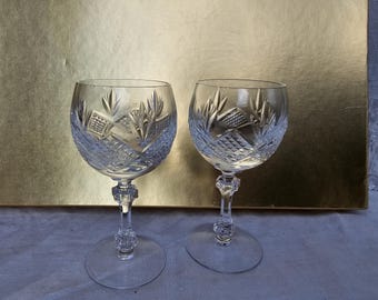 May include: Two clear crystal wine glasses with intricate cut glass designs. The bowls feature a pattern of leaves and geometric shapes. The stems have a decorative, textured design, and the bases are round. The background is a gold-colored surface.