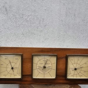 May include: A vintage wooden weather station with three square gauges. The gauges have a cream-colored background with black markings and hands. The wooden frame is a warm brown color, and the gauges are framed in a gold-colored metal.
