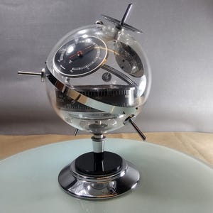 May include: A clear, spherical weather station with a black dial barometer and a silver base. The glass globe features a thermometer and a compass. The base is chrome with a black center. The design is retro and futuristic.