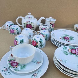 Może przedstawiać: Biały porcelanowy zestaw do herbaty z kwiatowym wzorem. Zestaw zawiera czajniczek, filiżanki, spodki, talerze, śmietankę i cukiernicę. Kwiatowy wzór przedstawia różowe i czerwone kwiaty z zielonymi liśćmi.