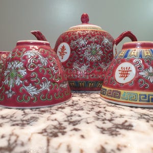 Op de afbeelding: Een rode en witte Chinese theeset met bloemenmotieven en Chinese karakters. De set bevat een theepot en vier kopjes.