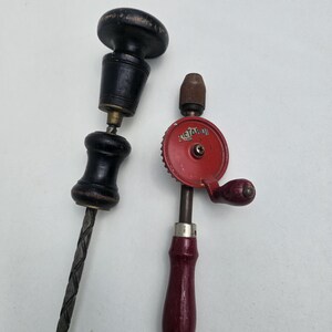 Peut inclure: Deux outils à main anciens : une tarière en bois noir et une perceuse à main rouge et argentée. La perceuse à main a une poignée rouge et un mécanisme à engrenages. La tarière a une poignée en bois foncé et une mèche torsadée en métal.