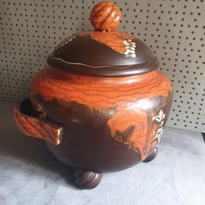 Può includere: Un grande vaso in ceramica marrone e arancione con un coperchio e un pomo attorcigliato. Il vaso ha un design marmoreo unico e tre piedi.
