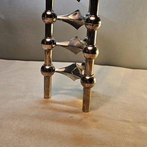 May include: A vintage silver and gold-toned candle holder. The candle holder has a vertical design with spherical accents and triangular, blade-like elements. The base has two cylindrical legs. The overall design is symmetrical and decorative.