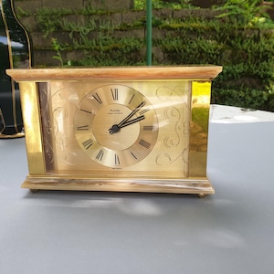 May include: A rectangular gold-toned clock with a light wood top and base. The clock face features Roman numerals and decorative scrollwork. The clock reads "Rando high-quartz".