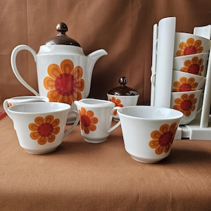 Könnte beinhalten: Vintage weißes Keramik-Teeservice mit retro-orangem und rotem Blumendesign. Das Set beinhaltet eine Teekanne, eine Zuckerdose, einen Sahnekännchen und mehrere Tassen. Ein weißer Tassenhalter zeigt zusätzliche Tassen.