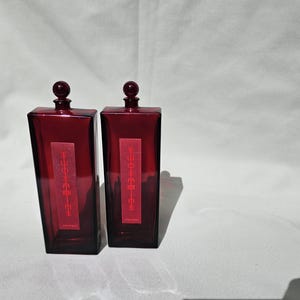 Könnte beinhalten: Zwei rechteckige, dunkelrote Glasflaschen mit abgerundeten Stöpseln. Jede Flasche hat ein rotes Etikett mit japanischem Text. Die Flaschen stehen auf einem weißen Hintergrund mit Schatten.