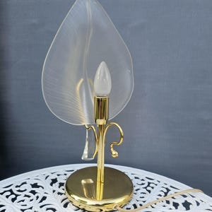 Könnte beinhalten: Eine Tischleuchte mit einem goldfarbenen, runden Sockel und einem schlanken Ständer mit geschwungenen Elementen. Eine einzelne Glühbirne sitzt in einer goldenen Fassung vor einem großen, halbtransparenten, mattierten Glasschirm in Blattform.