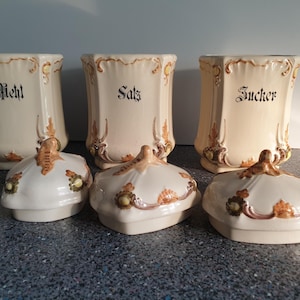Pode incluir: Três potes de cerâmica vintage com tampas, decorados com desenhos florais e detalhes dourados. Os potes estão etiquetados com "Mehl", "Salz" e "Zucker".