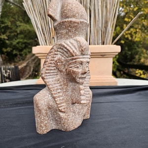 May include: A detailed, reddish-brown bust of an Egyptian pharaoh. The sculpture features a detailed headdress, facial features, and a stylized beard. The bust is made of a speckled stone material and is set against a natural background.