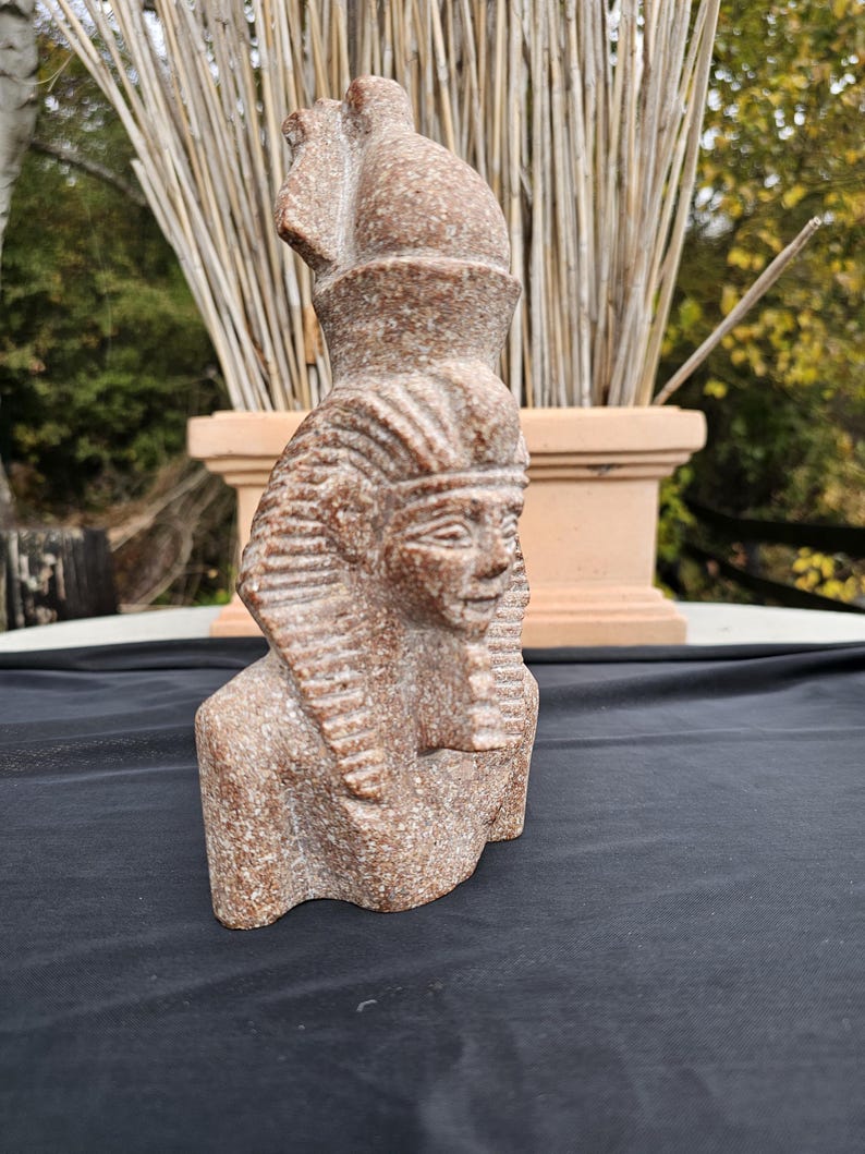 May include: A detailed, reddish-brown bust of an Egyptian pharaoh. The sculpture features a detailed headdress, facial features, and a stylized beard. The bust is made of a speckled stone material and is set against a natural background.