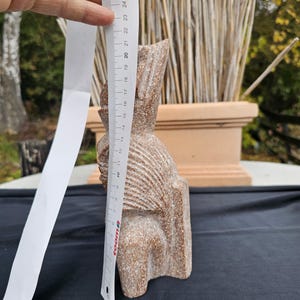 May include: A carved stone sculpture of a seated figure, approximately 9 inches tall. The sculpture is a light brown color with a textured surface. The figure has a detailed head and body, with carved lines. A ruler is held next to the sculpture.
