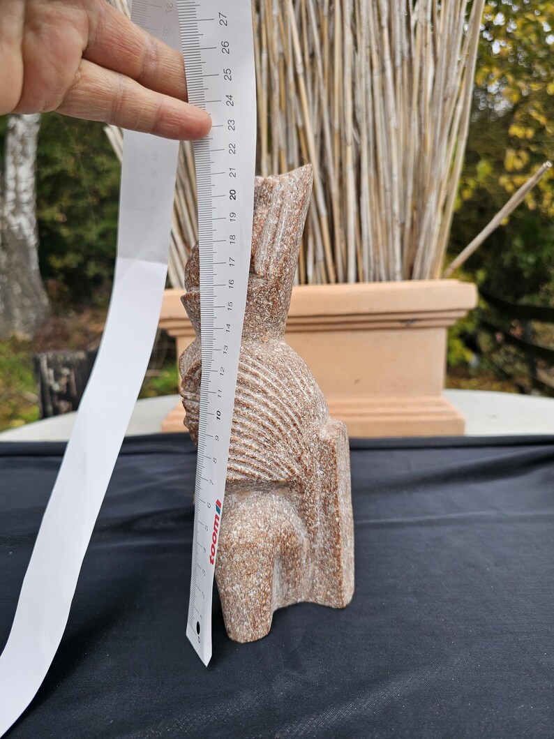 May include: A carved stone sculpture of a seated figure, approximately 9 inches tall. The sculpture is a light brown color with a textured surface. The figure has a detailed head and body, with carved lines. A ruler is held next to the sculpture.