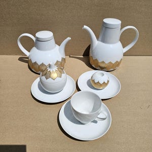 May include: A white porcelain tea set featuring two teapots, a lidded sugar bowl, a small spherical container, and a teacup with a saucer. Each piece is decorated with a repeating gold leaf-shaped pattern around the base or lower section.
