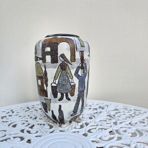 May include: A ceramic vase with a white base featuring a textured, multi-colored graphic illustration of figures carrying buckets, stylized buildings, and abstract shapes in shades of brown, olive green, blue, and black with a dark rim.