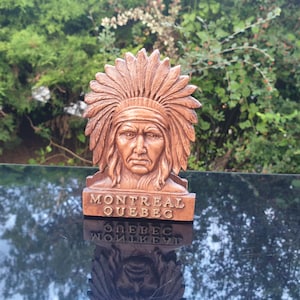 Può includere: Una statuetta di legno di un uomo nativo americano con un copricapo. La statuetta è di colore marrone e ha un viso scolpito. La base della statuetta dice "Montreal Quebec".