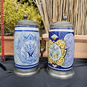 May include: Two ceramic beer steins with pewter lids. One stein features a blue and white floral design, while the other has a colorful floral and scroll design. Both steins have a cylindrical shape.