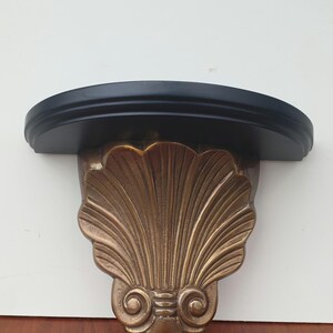 May include: A gold-colored, decorative wall shelf with a black top. The shelf is shaped like a seashell with intricate details.
