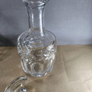 Könnte beinhalten: Eine klare Glas-Dekantierkaraffe mit dekorativem Verschluss. Die Karaffe hat einen breiten Boden und einen schmalen Hals. Der Körper der Karaffe ist mit einem sich wiederholenden Muster geätzt. Der Verschluss ist ebenfalls aus klarem Glas.