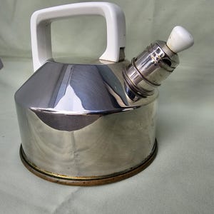 May include: A stainless steel stovetop kettle with a white plastic handle and spout knob. The kettle has a shiny, reflective surface and a circular base with a brass-colored rim. The handle is a large, rectangular shape.