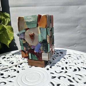 May include: A rectangular lamp featuring a mosaic exterior composed of various polished gemstones in shades of green, brown, purple, and translucent white. The lamp is mounted on a dark brown wooden base and includes a white power cord.
