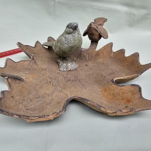 May include: A leaf-shaped dish with a bird and a flower sculpture. The dish is brown with a textured surface and a dark rim. The bird is gray and the flower is brown. A unique home decor item.