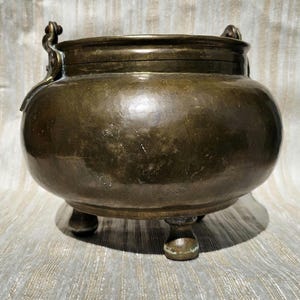 Pode incluir: Um pote de metal antigo, cor de bronze, com corpo arredondado e três pernas curtas. O pote tem duas alças ornamentadas e uma tampa. A superfície mostra sinais de idade e desgaste, com uma textura martelada.